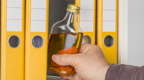 Alkohol am Arbeitsplatz Eine Flasche Alkohol wird von einer Hand zwischen Aktenordnern aus einem Regal herausgenommen.