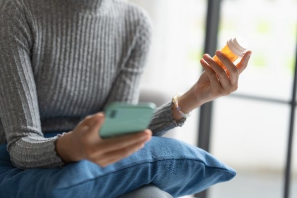 Suchtprävention Eine Frau hält in der einen Hand ein Smartphone, in der anderen eine Dose mit Medikamenten.