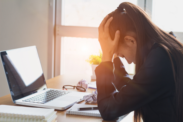 Eine Frau sitzt vor ihrem Laptop am Schreibtisch und stützt den Kopf in ihre Hände - sie wirkt überfordert und gestresst.